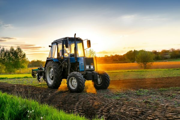 Modern blue tractor machinery plowing agricultural field meadow at farm at spring during sunset